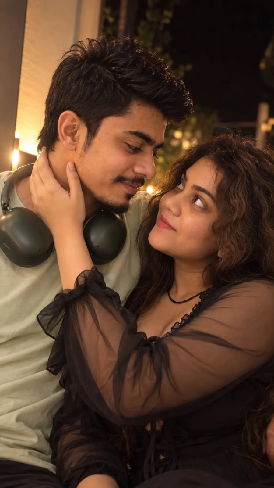 Manu and Jayu at night under warm string lights; her hand on his cheek, she looks up at him and smiles while he looks down at her.